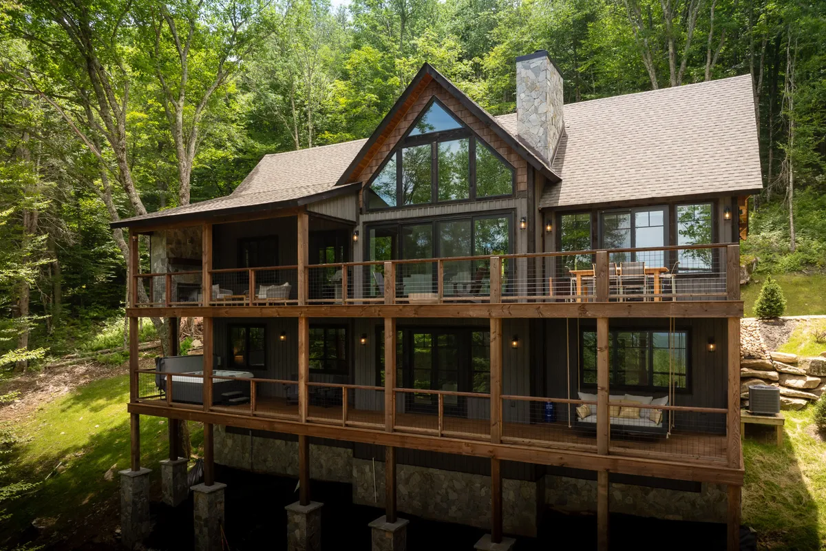 The image showcases a stunning mountain home exterior with expansive windows, a spacious wooden deck, a striking stone chimney, and is framed by lush, towering trees. The deck is positioned to enjoy panoramic sunset views and is set up for relaxation and gatherings in a tranquil, natural setting.