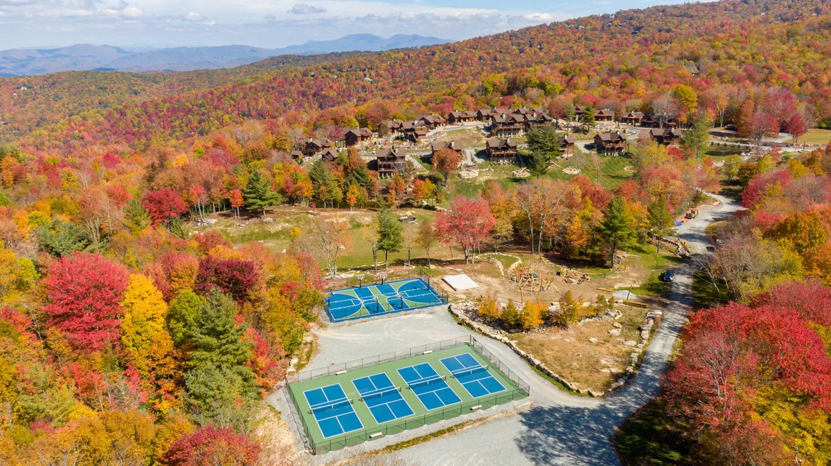 A vibrant scene of tennis courts nestled among colorful autumn foliage with residential cabins and a majestic mountain backdrop. The image radiates energy and activity, suggesting a community alive with sports, gatherings, and seasonal changes.