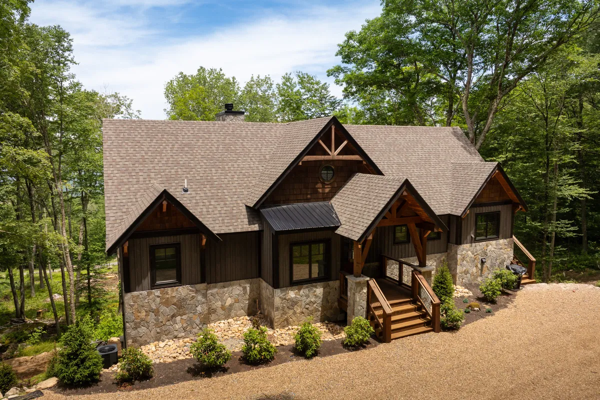 A welcoming exterior view of the mountain retreat, featuring a stone facade with rustic wooden beams, a landscaped path leading to the front porch, and the surrounding natural environment. The scene is bathed in warm, inviting light, hinting at sunset and the peaceful ambiance of the setting.