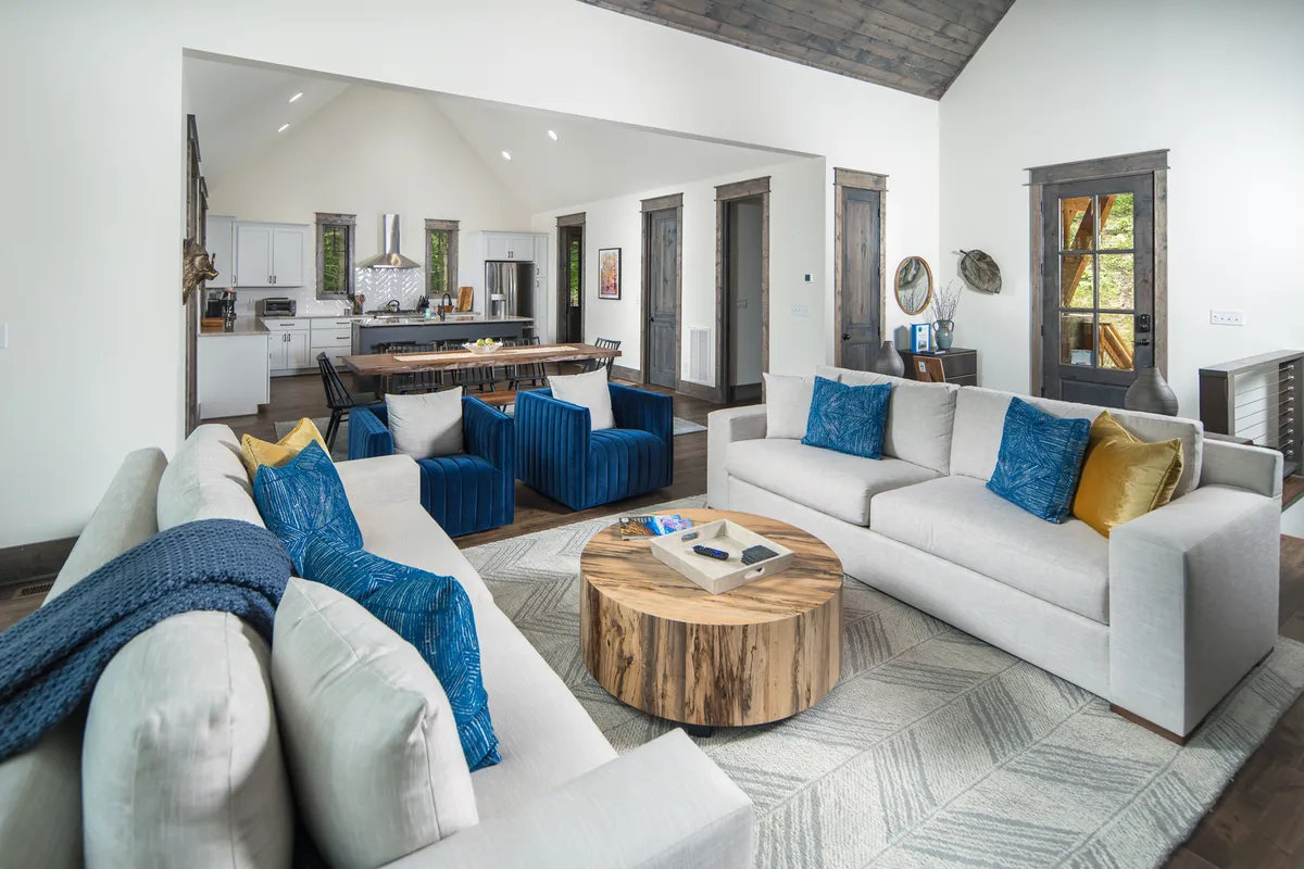 A welcoming seating arrangement featuring a plush sofa, round wooden coffee table, and blue accent chairs anchors the open-concept living space. In the background, a spacious kitchen with an island seamlessly connects to the living area, emphasizing the home's social layout.