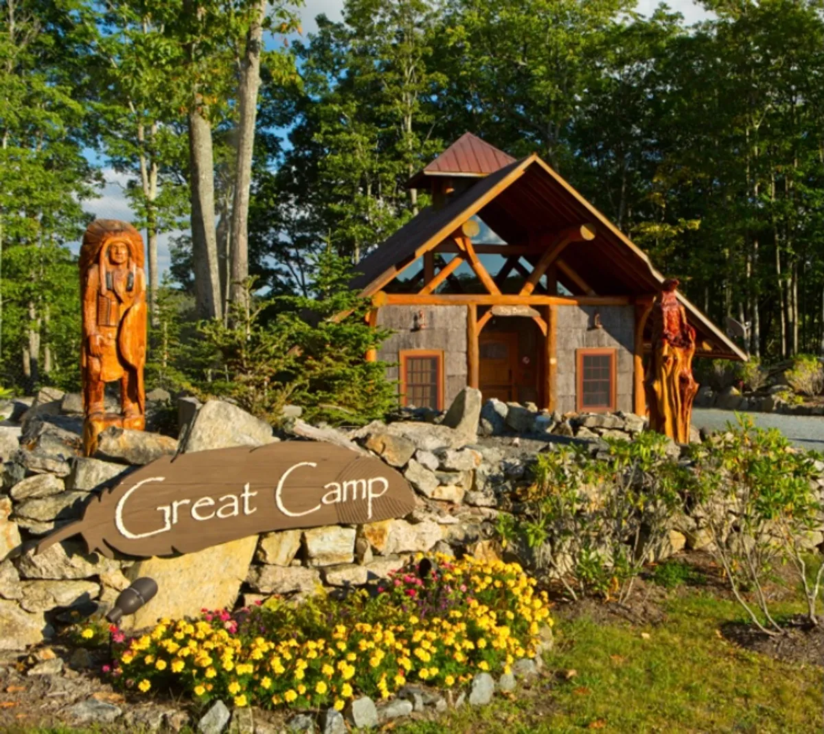 A rustic outdoor scene showing wooden sculptures, a stone wall, the 'Great Camp' sign, and a cabin structure surrounded by nature. The setting is inviting, ideal for family gatherings, outdoor activities, and storytelling by the campfire.