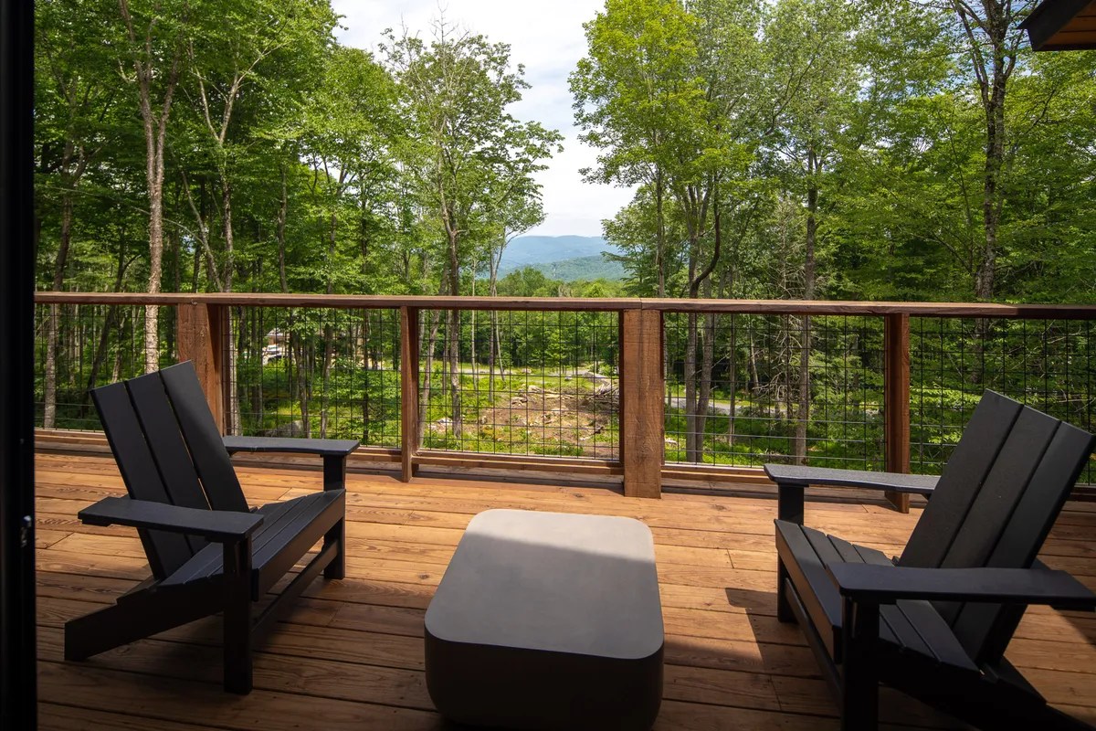 A spacious wooden deck overlooking a lush mountain landscape, with Adirondack chairs set up for relaxation. The view includes green trees and expansive sky, creating a sense of peace and openness. The deck’s easy access directly connects indoor comfort to outdoor adventure.
