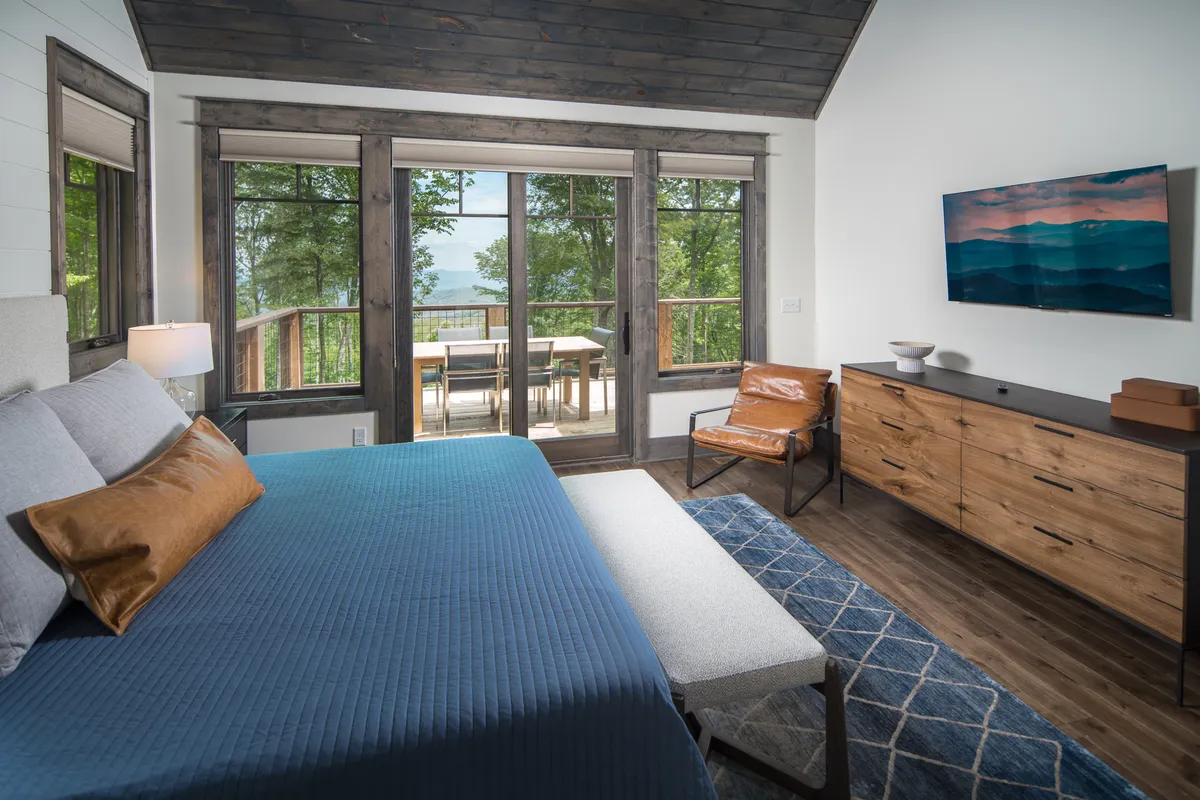 A spacious bedroom featuring a king-size bed with a blue quilt, large windows offering scenic views, a wooden dresser, and a comfortable armchair. Natural light floods the room, while the layout provides both comfort and privacy. The space exudes tranquility, perfect for restful nights.