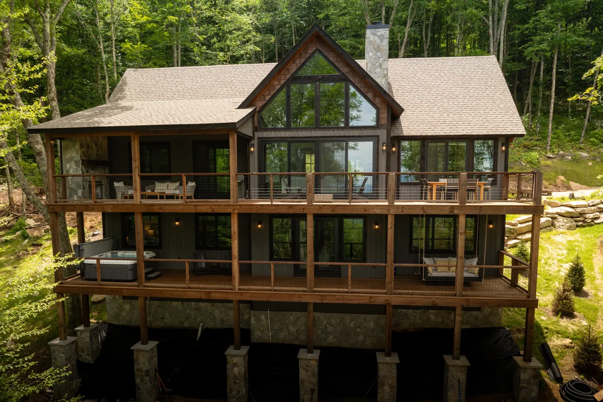An expansive exterior view featuring the home's large glass windows, wooden deck, and sturdy stone pillars, set amid surrounding trees. The deck area hints at outdoor gatherings, with the possibility of a fire pit and ample space for relaxation under the stars.