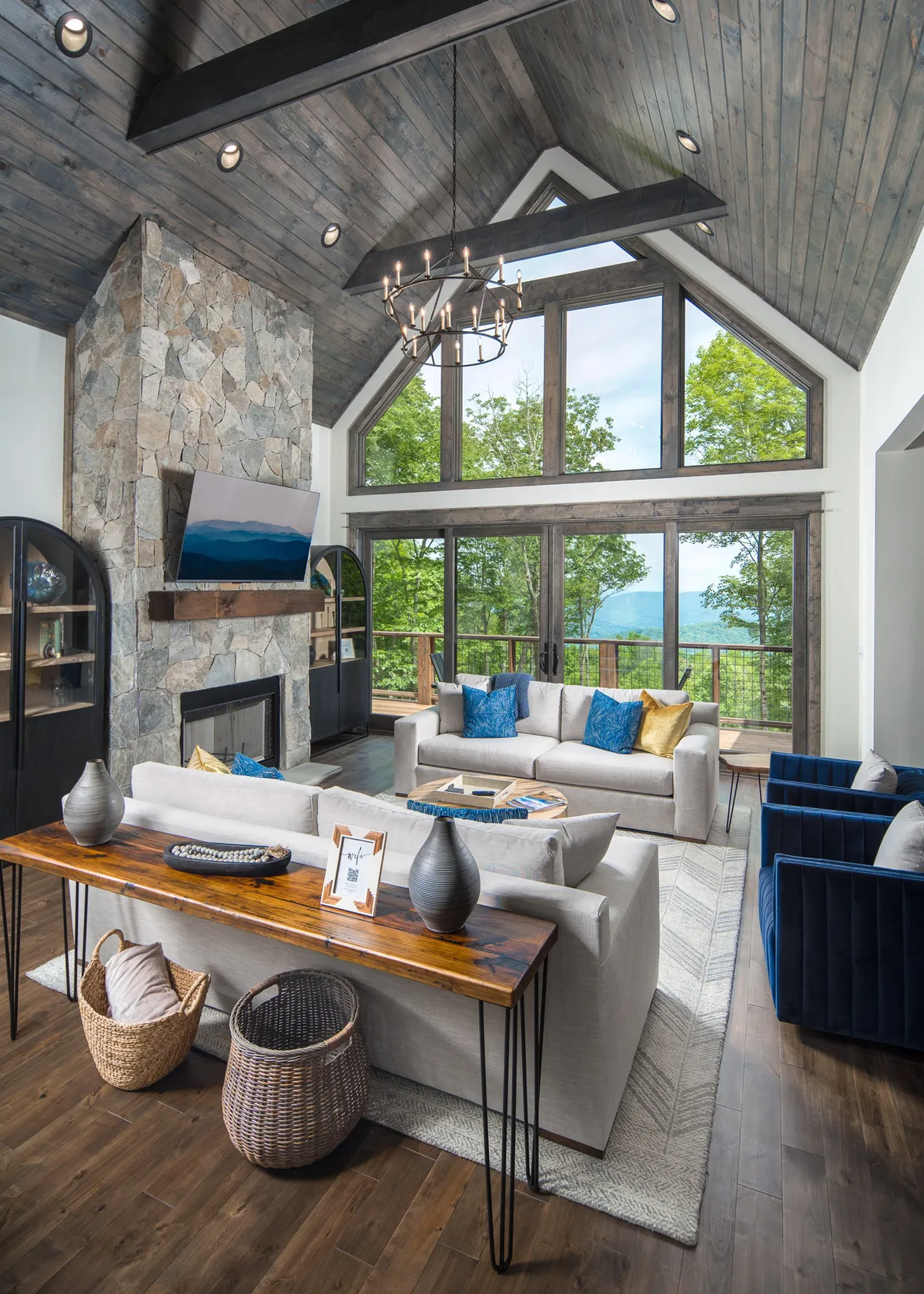 This image features a large stone fireplace as the centerpiece, set against expansive windows that flood the room with natural light. The cozy seating area with colorful cushions and a wooden coffee table invites relaxation and group gatherings. A chandelier overhead adds a touch of elegance to the inviting, spacious Great Room.