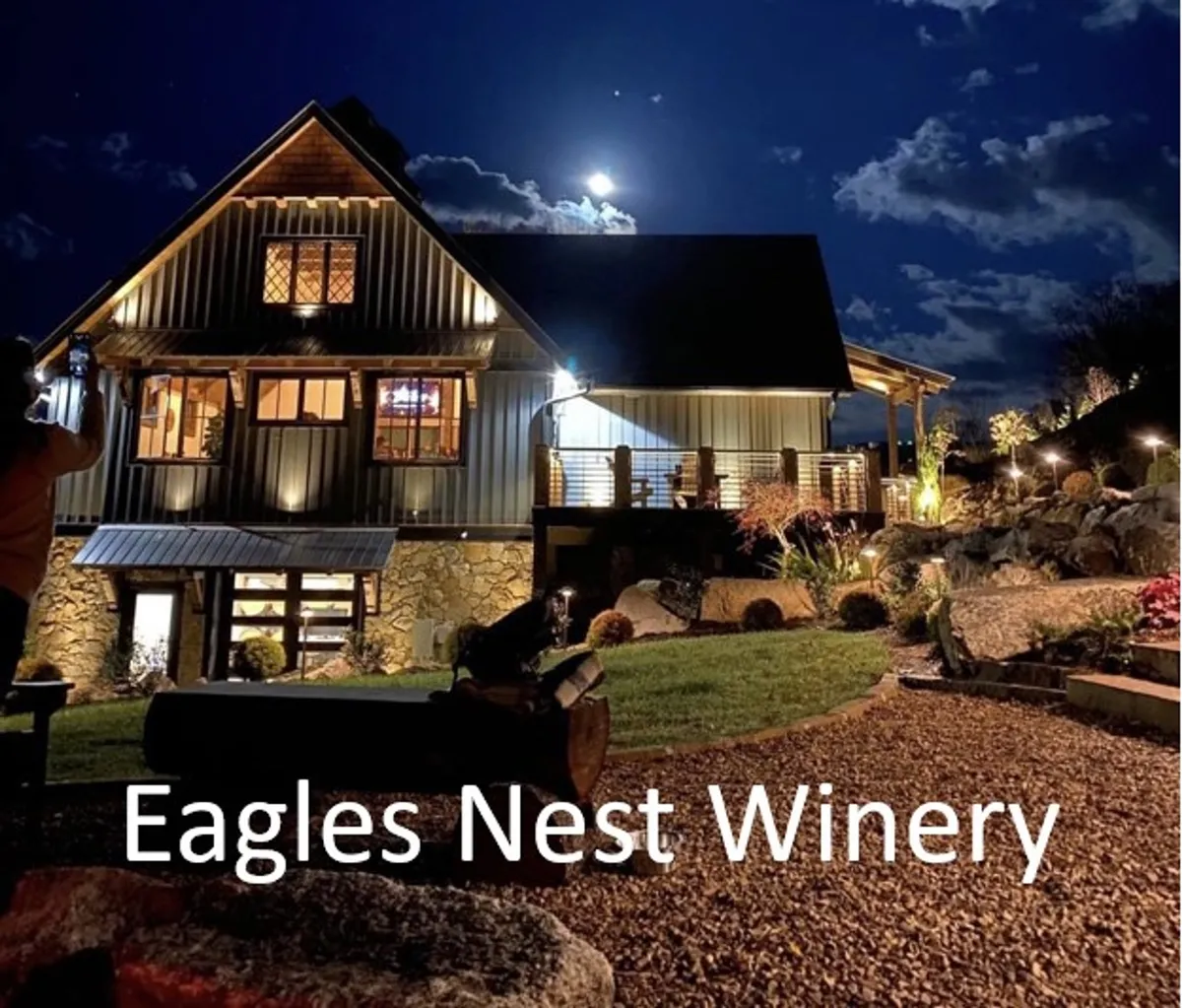 The image features the illuminated Eagles Nest Winery building at dusk, with a moonlit sky and a beautifully landscaped garden in the foreground. The warm glow from the winery contrasts with the cool night, creating an inviting atmosphere for evening gatherings and wine tasting.