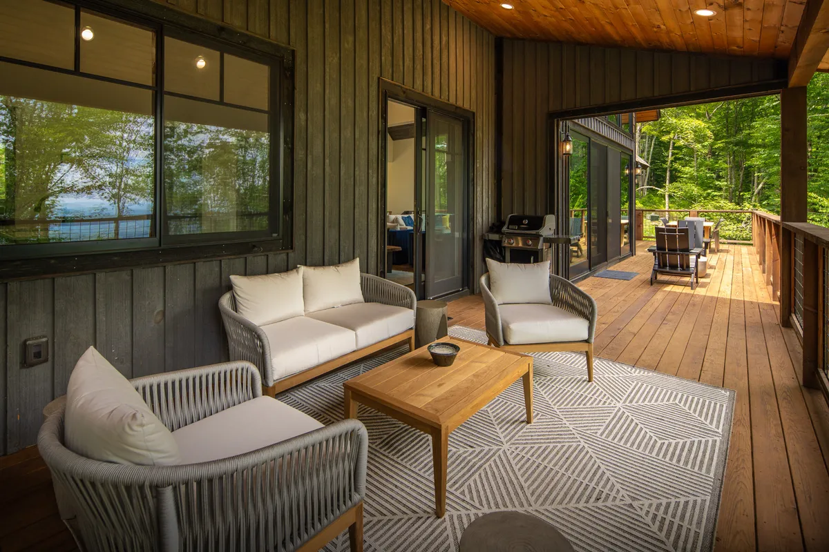 An exterior setting with a comfortable seating area, a wooden table, and a rug with a geometric pattern, surrounded by lush greenery. The tranquil atmosphere suggests a perfect spot for socializing, dining outdoors, or simply watching pets play in a natural, secure environment.