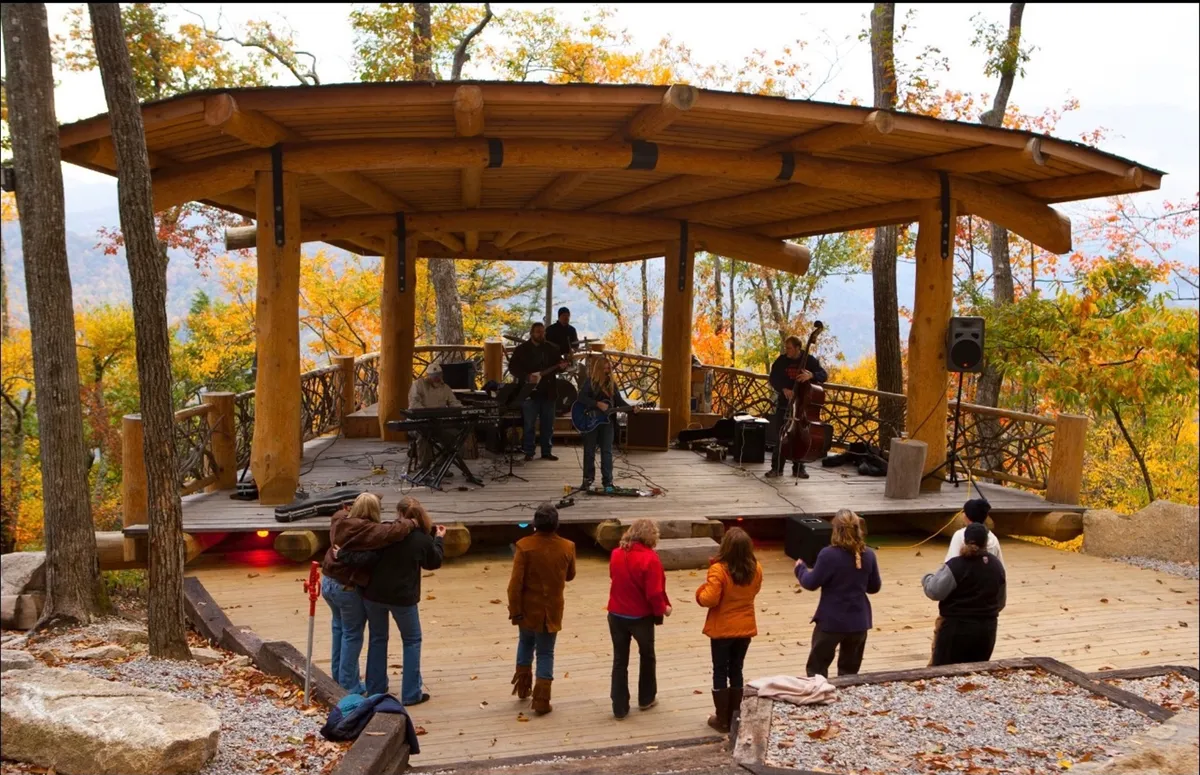 An outdoor amphitheater set amidst natural surroundings, with musicians on stage and audience members enjoying live music. The scene conveys a sense of lively community, entertainment, and shared joy in a mountain setting.